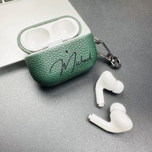 May include: Green textured leather AirPod case with the name "Michael" in cursive script. The case is open, revealing the white interior. Two white wireless earbuds are next to the case. A silver carabiner clip is attached to the case.