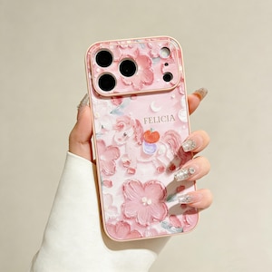 May include: Pink phone case with a floral and unicorn design. The case features the name "FELICIA" and a painted style with a light pink background and darker pink flowers. The unicorn has a red apple on its back.