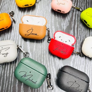 May include: A collection of colorful, personalized AirPod cases. The cases are in various colors, including brown, red, green, and black, with names written in cursive. Each case has a silver carabiner clip attached.