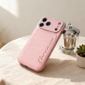 Puede incluir: Una funda de teléfono rosa con un patrón floral en relieve y el nombre "Lorenzo" en letras doradas. La funda tiene un protector de lente transparente y un borde metálico. La funda está sobre una superficie color crema.