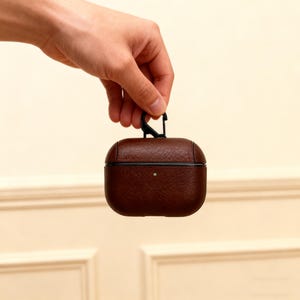 May include: A brown leather AirPods Pro case with a black carabiner clip. The case has a textured surface and a small silver dot on the front. The case is held by a hand against a neutral background.