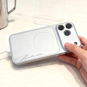 Puede incluir: Funda plateada para teléfono con un círculo de carga inalámbrica y tres lentes de cámara. La funda tiene el nombre "Isabella" en escritura cursiva. Un cable de carga blanco está conectado.