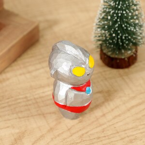 May include: A small, silver-colored figurine of a cartoon character with yellow eyes, a red collar, and a blue accent. The figurine is standing on a wooden surface, with a small Christmas tree in the background.