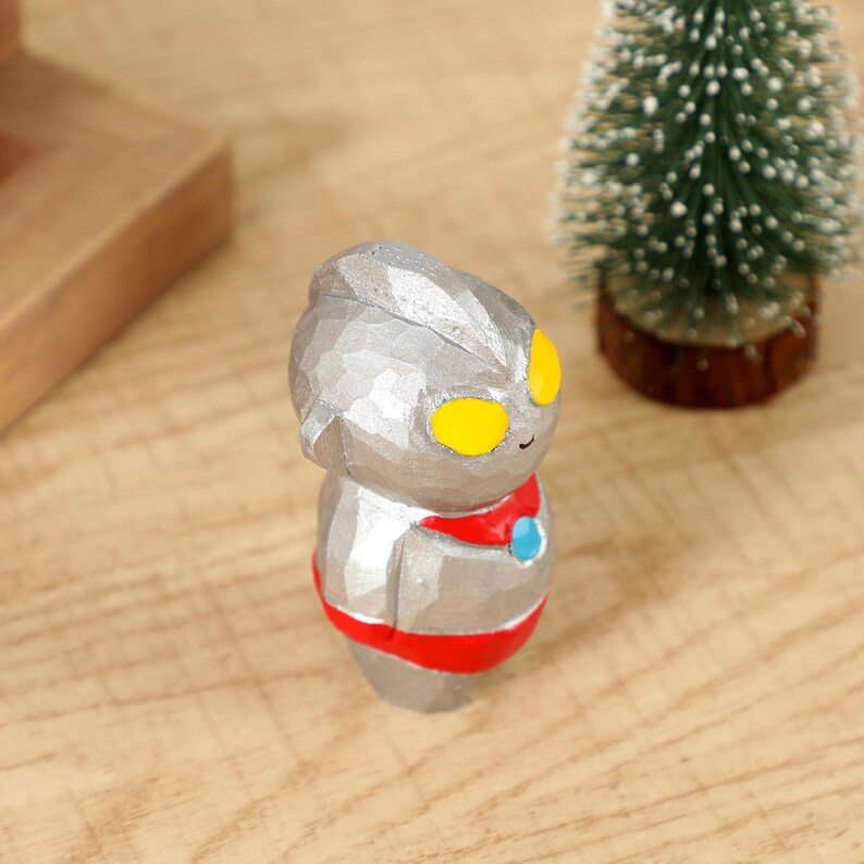 May include: A small, silver-colored figurine of a cartoon character with yellow eyes, a red collar, and a blue accent. The figurine is standing on a wooden surface, with a small Christmas tree in the background.