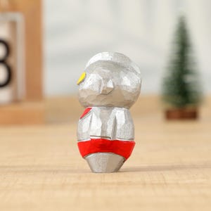 May include: A small, carved wooden figurine of a character with a silver body, red belt, and yellow eye details. The figure has a textured surface and stands on a wooden surface. A small green Christmas tree is in the background.