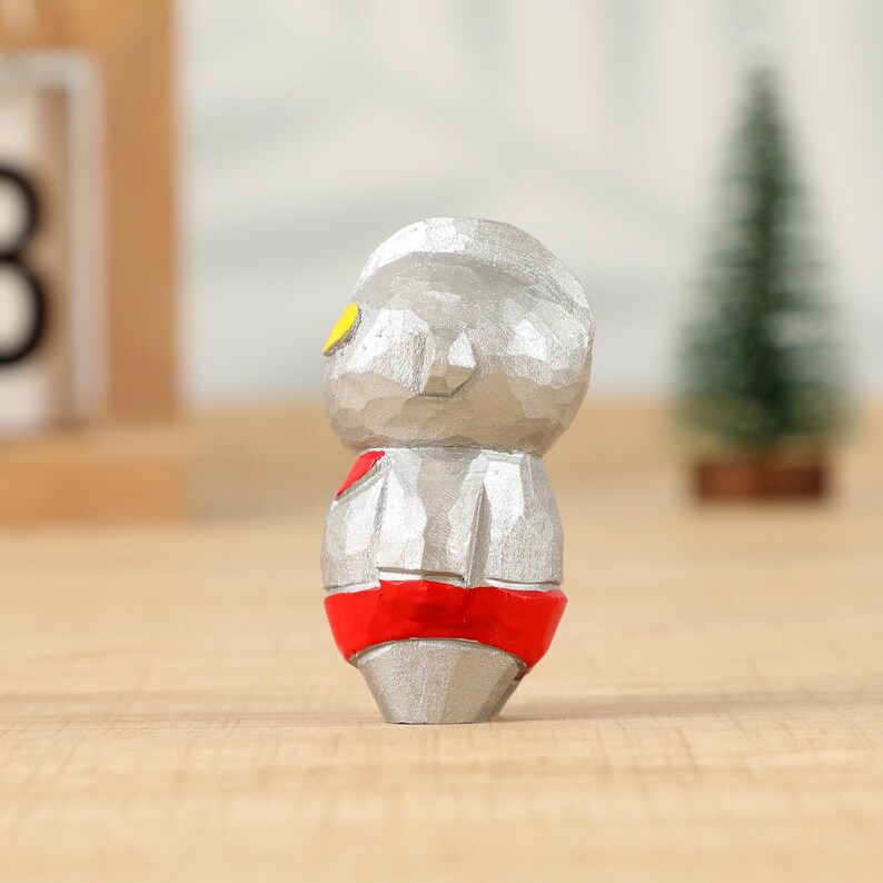May include: A small, carved wooden figurine of a character with a silver body, red belt, and yellow eye details. The figure has a textured surface and stands on a wooden surface. A small green Christmas tree is in the background.
