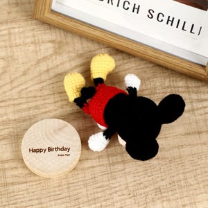 May include: A handmade crochet Mickey Mouse toy in red, black, yellow, and white, lying on a wooden surface. A wooden disc with the text "Happy Birthday From Yori" is next to the toy. A framed print with the text "RICH SCHILLI" is in the background.