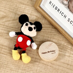 May include: A handmade crochet Mickey Mouse doll in classic colors: black ears, red shorts with white buttons, yellow shoes, and white gloves. A wooden disc with "Happy Birthday" text is next to the doll.