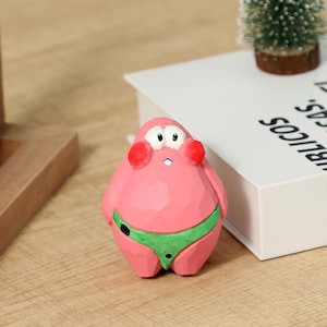 May include: A pink, wooden figurine of a cartoon character with a round body, white eyes, and red cheeks. The character wears a green swimsuit. The figurine is on a wooden surface.