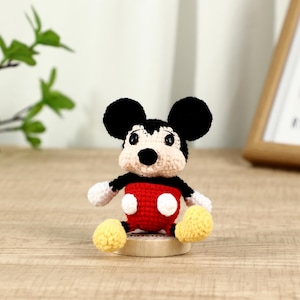 May include: A handmade crochet Mickey Mouse doll. The doll has black ears, a black and white face, a red body with white buttons, white gloves, and yellow shoes. It is sitting on a small wooden base.
