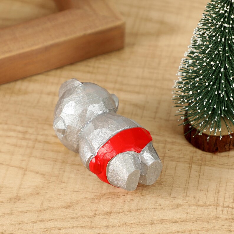 May include: A silver-colored bear figurine with a red band around its waist. The bear is lying down on a wooden surface. A small green Christmas tree is in the background.