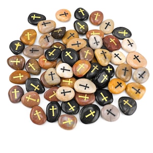 May include: A collection of smooth, rounded stones in various colors, including black, brown, tan, and white. Each stone features a gold or black cross symbol. The stones are scattered, creating a natural and textured appearance.