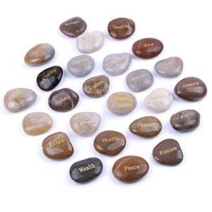 May include: A selection of polished stones in shades of brown, grey, and beige. Each stone features a gold inscription with words such as "Love", "Peace", "Health", and "Joy". The stones are arranged on a white surface.