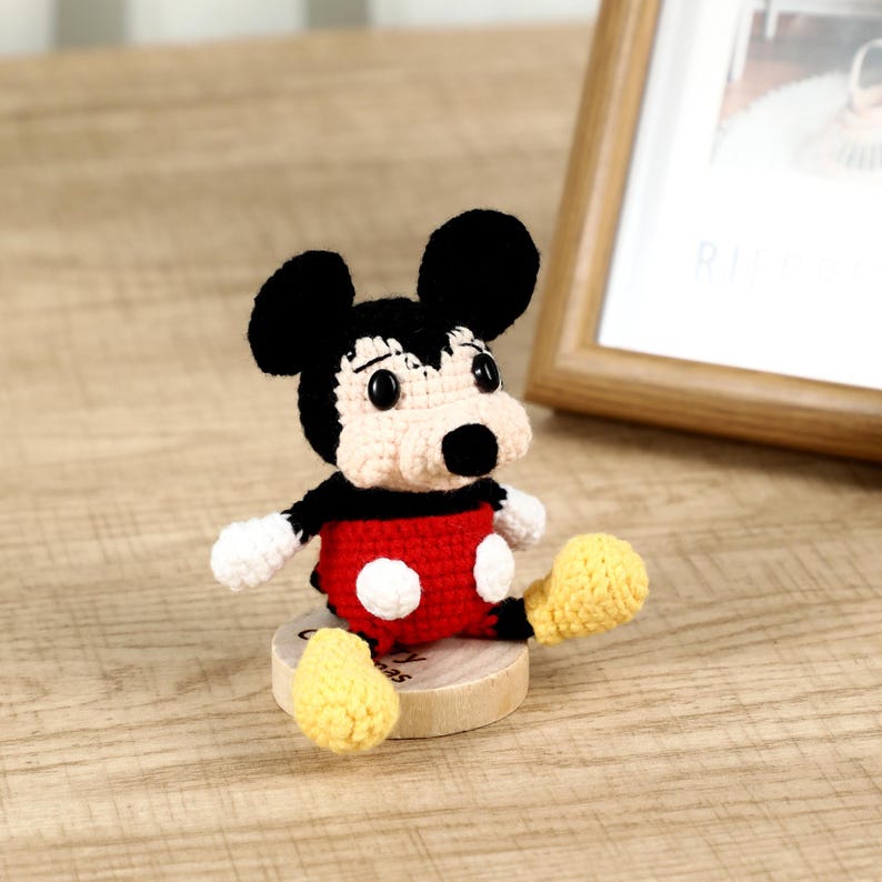 May include: A handmade crochet Mickey Mouse figure. The figure has black ears, a white face, and a red body with white buttons. The figure is sitting on a small wooden base. The figure's feet are yellow.