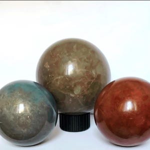 May include: Three polished stone spheres in shades of green, brown, and red. The largest sphere is brown and rests on a black base. The other two spheres are a teal green and a deep red. The spheres are smooth and reflective.