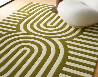 Puede incluir: Una alfombra moderna con un diseño geométrico en verde oliva y crema. La alfombra presenta líneas curvas y rectas, creando un patrón abstracto. Un sillón blanco y una mesa auxiliar con una escultura están en el fondo.