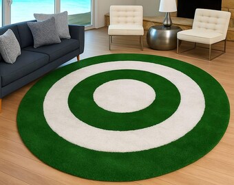 May include: A round rug with a green and white target design. The rug features a green outer ring, a white middle ring, and a white center. The rug is placed on a hardwood floor in a living room setting.