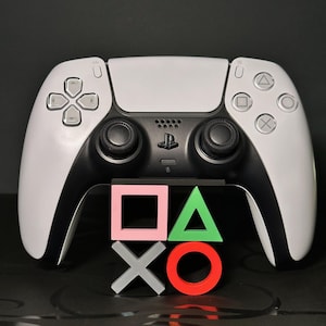 May include: A white and black PlayStation 5 controller with a set of colourful button symbols in front. The controller has a black centre section with the PlayStation logo and white side panels. The button symbols are pink, green, grey, and red.