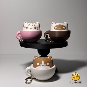 Puede incluir: Tres figuras con forma de gato en tazas de té, cada una con un diseño diferente, se exhiben en un soporte negro. Las tazas son rosas, marrones y blancas. Cada taza tiene una cadena plateada. Las figuras tienen expresiones faciales variadas.