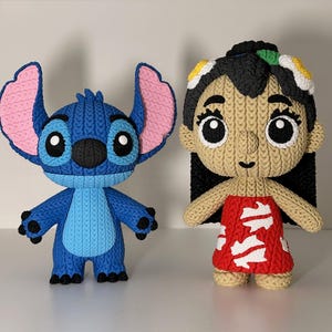Puede incluir: Dos figuras de personajes hechas a mano y tejidas. Una es azul con grandes orejas rosas, que se asemeja a un extraterrestre de dibujos animados. La otra es una ni&ntilde;a con un vestido rojo con detalles blancos, cabello negro y una diadema floral. Ambas figuras tienen ojos grandes y expresivos.