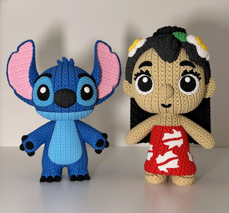 Puede incluir: Dos figuras de personajes hechas a mano y tejidas. Una es azul con grandes orejas rosas, que se asemeja a un extraterrestre de dibujos animados. La otra es una ni&ntilde;a con un vestido rojo con detalles blancos, cabello negro y una diadema floral. Ambas figuras tienen ojos grandes y expresivos.