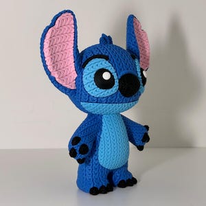 Puede incluir: Figura azul de Stitch, personaje de la pel&iacute;cula Lilo & Stitch. Tiene grandes orejas con interior rosa, ojos negros y aspecto de punto. La figura est&aacute; sobre una superficie blanca. Un objeto de colecci&oacute;n.
