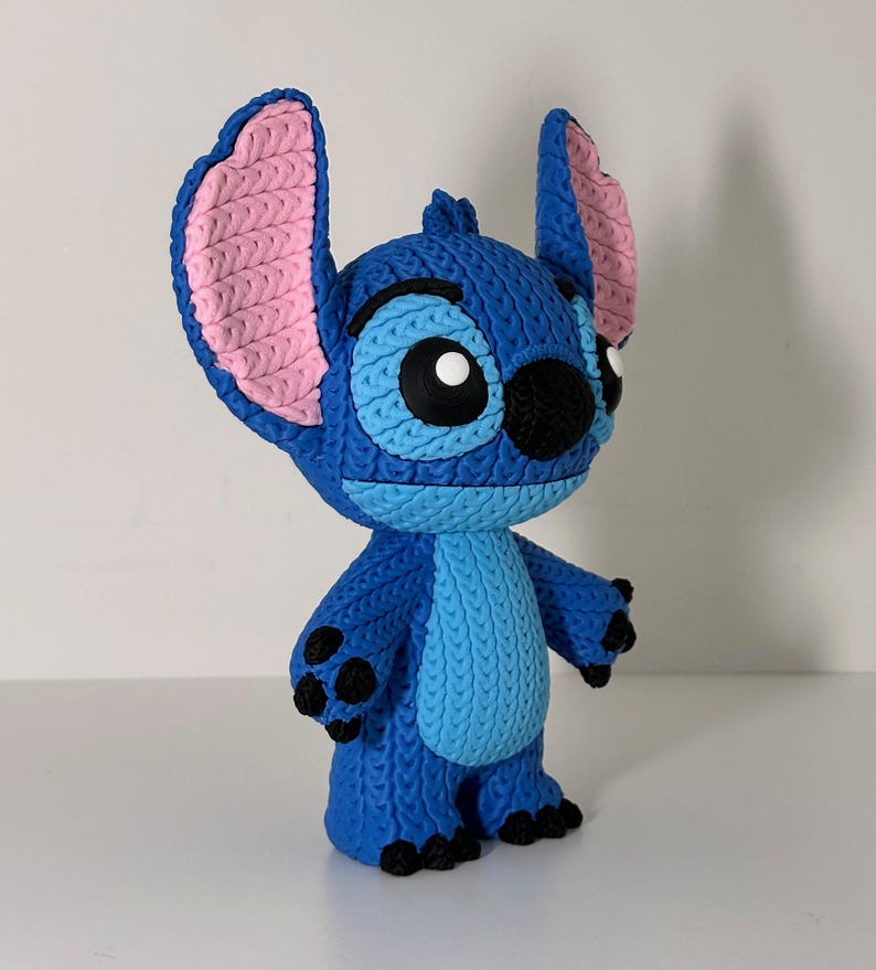 Puede incluir: Figura azul de Stitch, personaje de la pel&iacute;cula Lilo & Stitch. Tiene grandes orejas con interior rosa, ojos negros y aspecto de punto. La figura est&aacute; sobre una superficie blanca. Un objeto de colecci&oacute;n.
