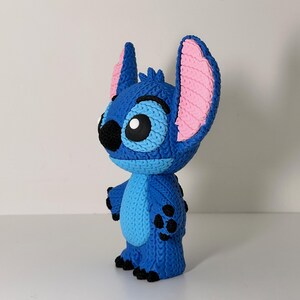 Puede incluir: Figura azul del personaje Stitch de la pel&iacute;cula animada Lilo & Stitch. La figura tiene orejas grandes con interiores rosas, una nariz negra y ojos grandes. La figura est&aacute; de pie sobre una superficie blanca.
