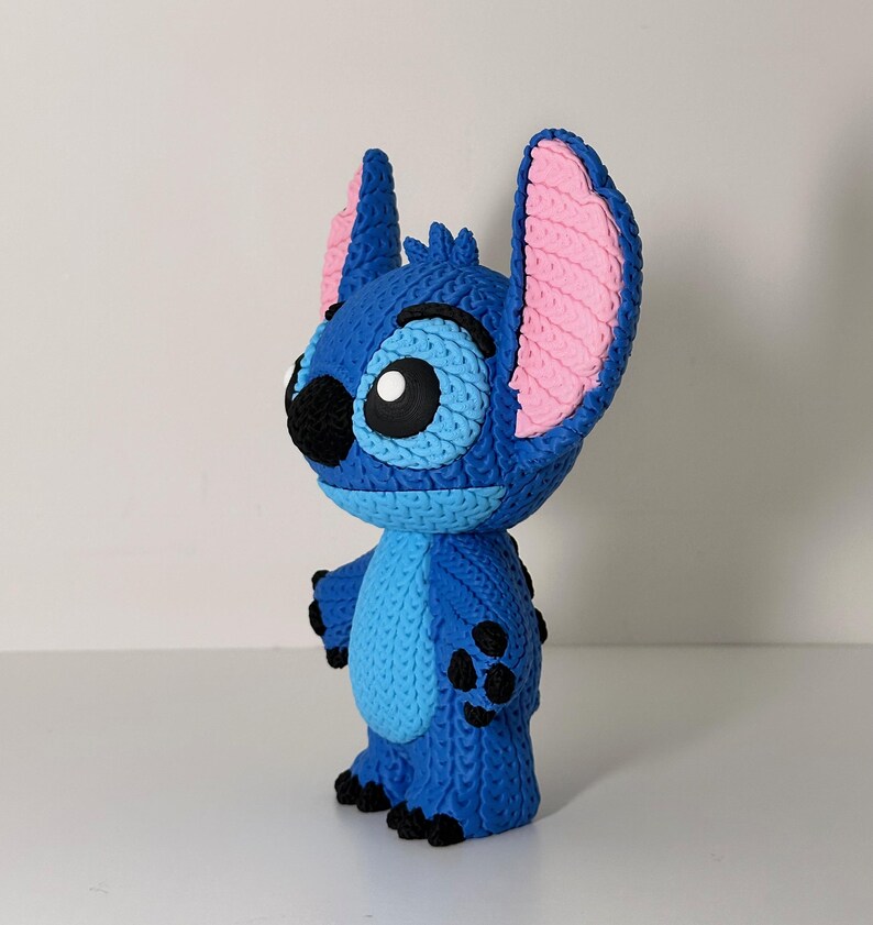 Puede incluir: Figura azul del personaje Stitch de la pel&iacute;cula animada Lilo & Stitch. La figura tiene orejas grandes con interiores rosas, una nariz negra y ojos grandes. La figura est&aacute; de pie sobre una superficie blanca.