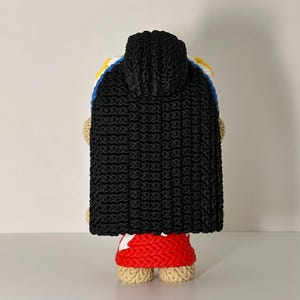 Puede incluir: Mu&ntilde;eca de ganchillo hecha a mano, vista desde atr&aacute;s, con una capa con capucha negra. La mu&ntilde;eca tiene una falda roja y piernas color canela. La capa est&aacute; sobre un dise&ntilde;o blanco, azul y amarillo.