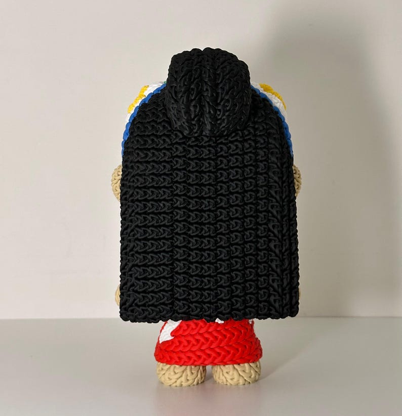Puede incluir: Mu&ntilde;eca de ganchillo hecha a mano, vista desde atr&aacute;s, con una capa con capucha negra. La mu&ntilde;eca tiene una falda roja y piernas color canela. La capa est&aacute; sobre un dise&ntilde;o blanco, azul y amarillo.