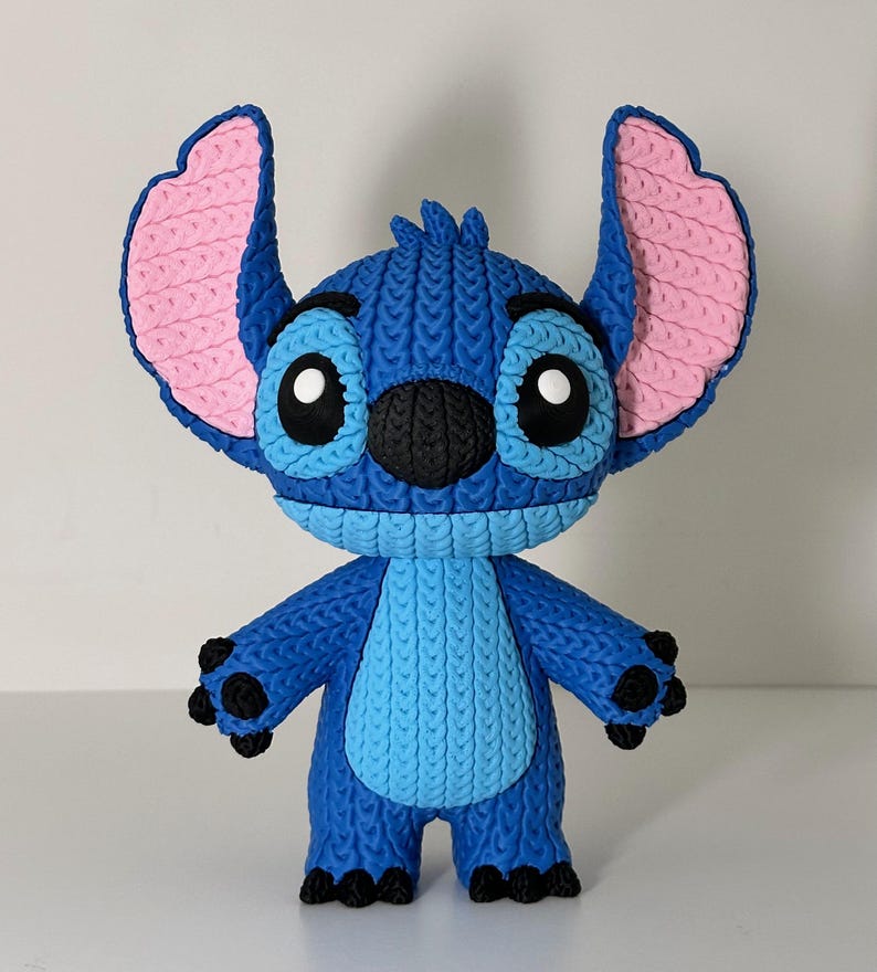 Puede incluir: Figura azul de Stitch con orejas grandes, ojos negros y nariz negra. La figura tiene un aspecto texturizado y tejido, con detalles rosas en las orejas y detalles negros en manos y pies. El personaje est&aacute; de pie.