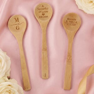 Puede incluir: Tres cucharas de madera con texto grabado. Una tiene las iniciales "M and G", otra dice "A Spoonful of Love E+K 2028", y la tercera "Bonnie & Wayne 10.12.2027". Las cucharas están dispuestas sobre una superficie rosa con rosas blancas.