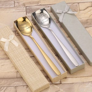 Peut inclure: Trois cuillères dans des coffrets cadeaux. Une cuillère est dorée avec le texte "Modesty's Spoon". Une autre cuillère est argentée avec le texte "Vicky's Kitchen". Les boîtes sont beiges et grises, avec des nœuds en ruban blanc.