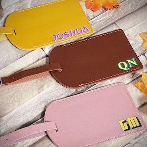 Puede incluir: Tres etiquetas de equipaje personalizadas en amarillo, marrón y rosa. La etiqueta amarilla tiene el nombre "JOSHUA" en morado. La etiqueta marrón tiene las iniciales "QN" en verde. La etiqueta rosa tiene las iniciales "GM" en amarillo y azul.