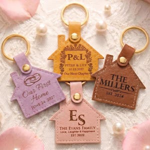Puede incluir: Cuatro llaveros con forma de casa en tonos morado, beige y rosa, con aros dorados. Cada llavero presenta texto personalizado, incluyendo nombres, fechas y detalles familiares. Los llaveros están diseñados para ocasiones especiales.