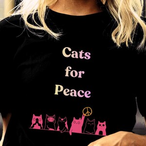 Può includere: T-shirt nera con la scritta "Cats for Peace" in un font sfumato. Sotto, una fila di illustrazioni di gatti rosa, uno dei quali tiene un simbolo della pace.