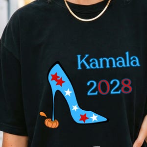 Könnte beinhalten: Schwarzes T-Shirt mit einem Design eines blauen High-Heel-Schuhs, verziert mit roten und weißen Sternen. Der Text "Kamala 2028" ist in Blau und Rot dargestellt. Ein kleiner orangefarbener Kürbis ist ebenfalls vorhanden.