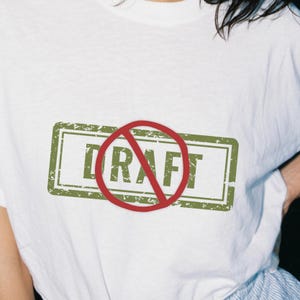May include: White t-shirt with a green rectangular stamp design featuring the word "DRAFT" in green, crossed out by a red circle. The shirt is made of a soft material.