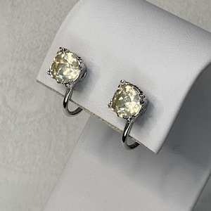 May include: A pair of clip-on earrings featuring pale yellow gemstones set in a silver-colored metal. The gemstones have a rounded, faceted shape, and the earrings are displayed on a white surface.