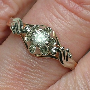 May include: A gold ring with a delicate, ornate design featuring a single, round, clear diamond in the center.