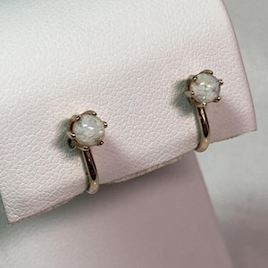 May include: A pair of gold-toned clip-on earrings. Each earring features a round, iridescent opal gemstone set in a pronged setting. The earrings are displayed on a white surface.