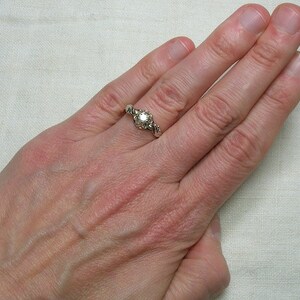 May include: A silver ring with a round, clear gemstone surrounded by smaller gemstones. The ring is on a person's finger.