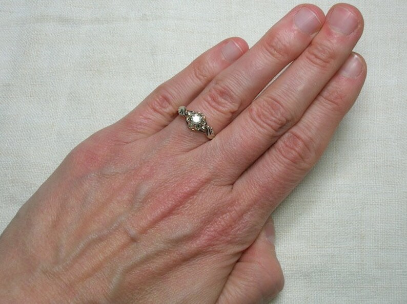 May include: A silver ring with a round, clear gemstone surrounded by smaller gemstones. The ring is on a person's finger.