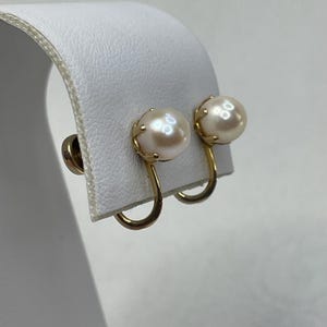 May include: A pair of vintage pearl earrings. Each earring features a round, creamy-white pearl set in a gold-tone metal frame. The earrings have a screw-back closure. The pearls have a subtle luster, and the gold setting adds a touch of elegance.