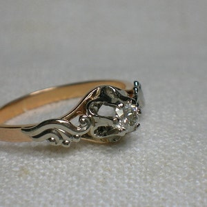 May include: A vintage engagement ring with a single round diamond set in a white gold setting. The band is made of rose gold and features a delicate, ornate design.