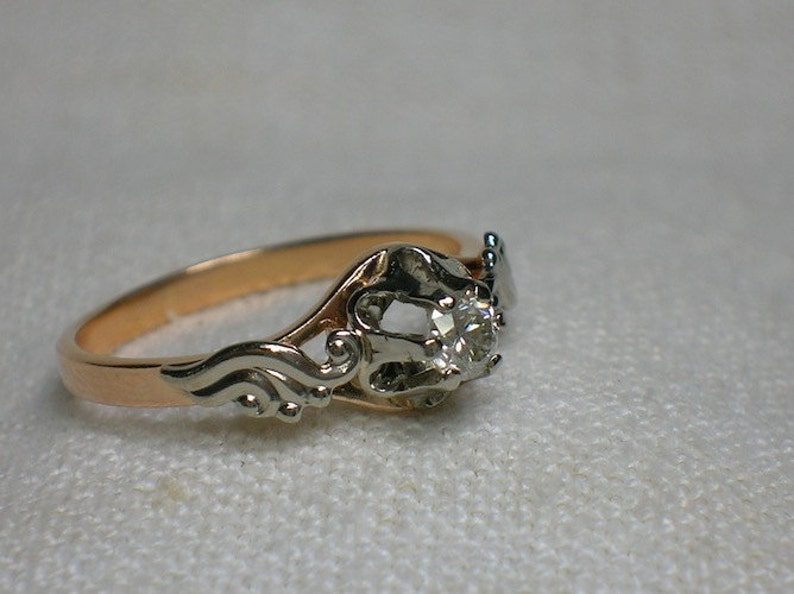 May include: A vintage engagement ring with a single round diamond set in a white gold setting. The band is made of rose gold and features a delicate, ornate design.