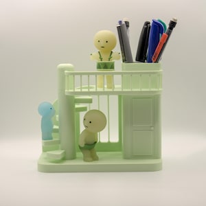 Könnte beinhalten: Ein hellgrüner Stifthalter in Form eines zweistöckigen Gebäudes mit Treppe und drei kleinen Figuren. Die obere Ebene enthält Stifte und Marker. Eine Figur ist hellblau, die anderen beiden sind hellgelb.