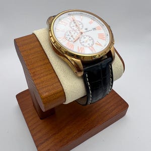 May include: A gold-toned wristwatch with a white face and rose-gold accents, featuring a black leather band with white stitching. The watch is displayed on a wooden stand with a cream-colored cushion.