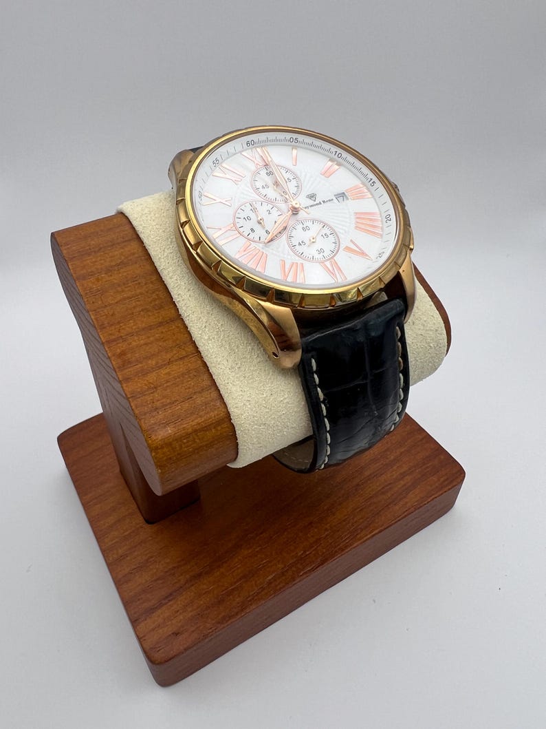 May include: A gold-toned wristwatch with a white face and rose-gold accents, featuring a black leather band with white stitching. The watch is displayed on a wooden stand with a cream-colored cushion.